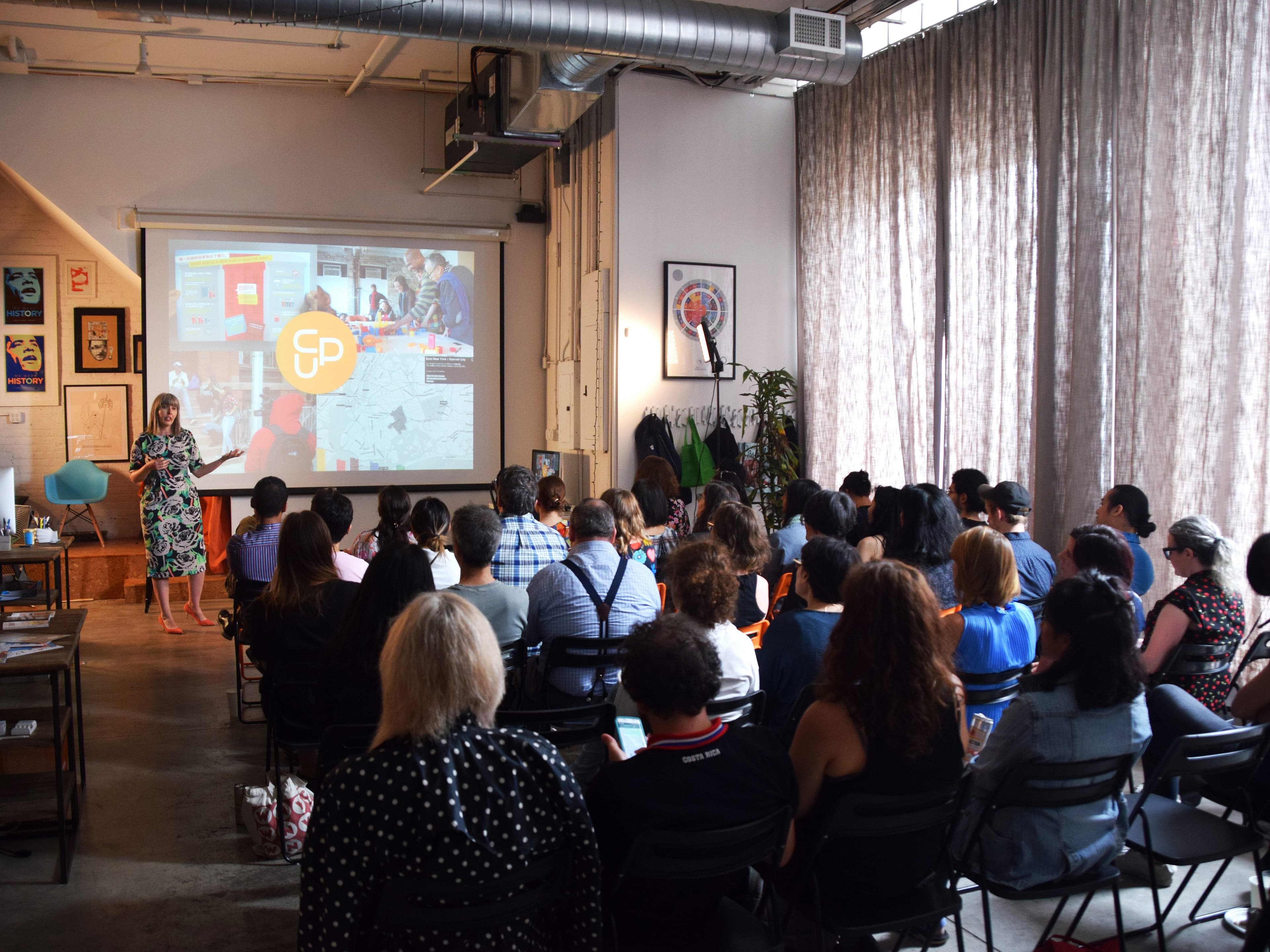 A woman in a floral dress is presenting to a seated audience in a modern, industrial-style room. A large screen behind her displays a colorful presentation. The attendees are focused on the speaker and some are taking notes. A tall plant and artwork adorn the room.