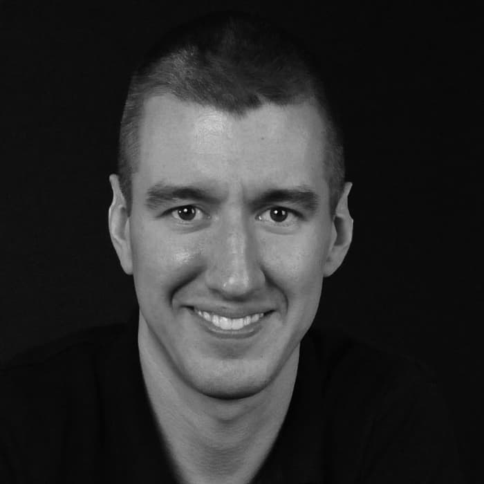 A black and white portrait of a person with short hair, wearing a dark shirt. The individual is smiling and looking directly at the camera against a plain dark background.