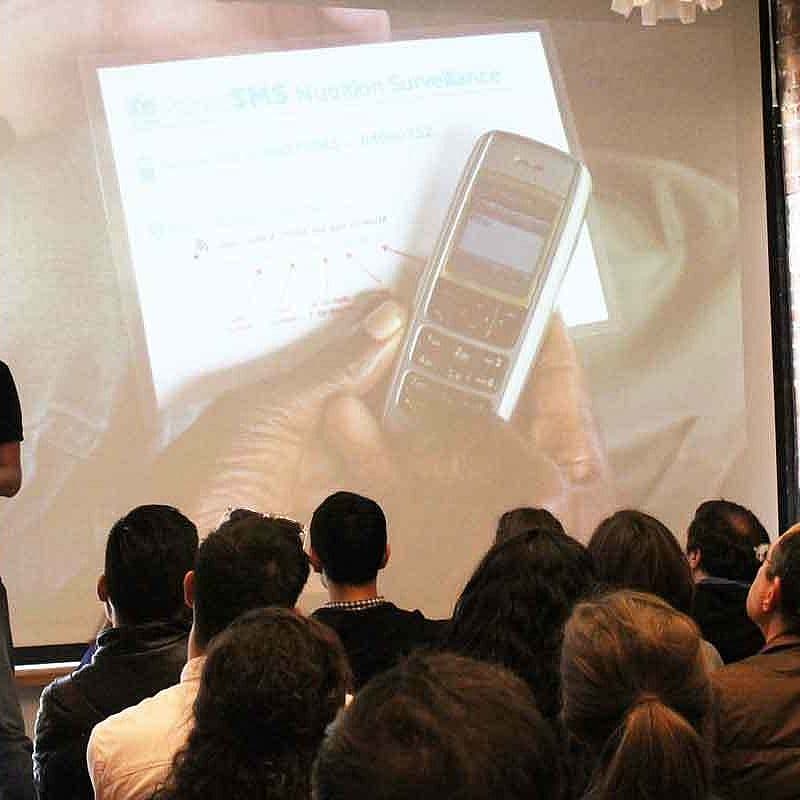 A man stands and presents to a seated audience. Behind him, a large projection screen displays an image of a hand holding an older model mobile phone, with text at the top reading "SMS Nutrition Surveillance." The room has brick walls and large windows.