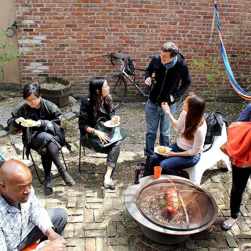 A group of people is gathered in an outdoor setting with a brick wall and hammocks in the background. They are seated and standing around a fire pit, enjoying food and engaging in conversation. The atmosphere appears relaxed and casual.
