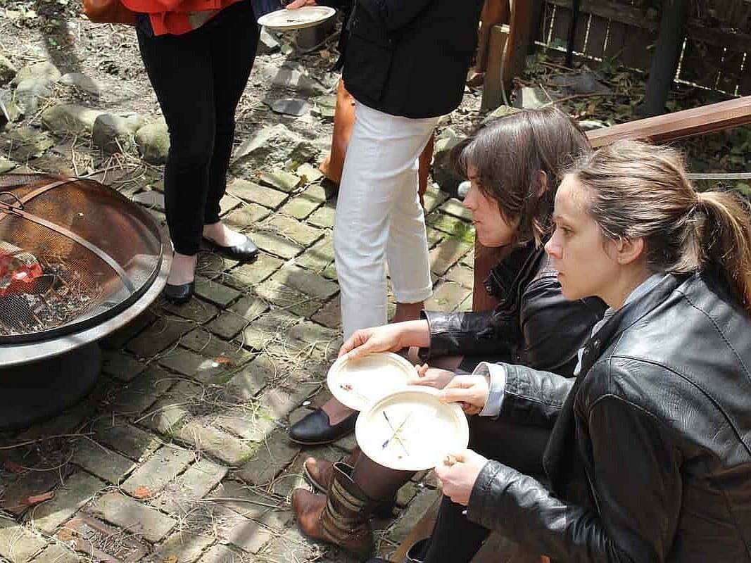 Two women with plates of food sit next to a fire pit with a piece of meat cooking on a rotisserie, surrounded by cobblestone ground. Other people in the background are standing and holding plates. The scene takes place outdoors, possibly in a garden or backyard.