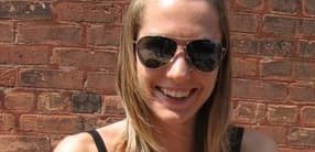 A person with long blonde hair is smiling and wearing dark sunglasses. They are standing in front of a brick wall and wearing a black tank top. The weather appears to be sunny based on the lighting and shadows.