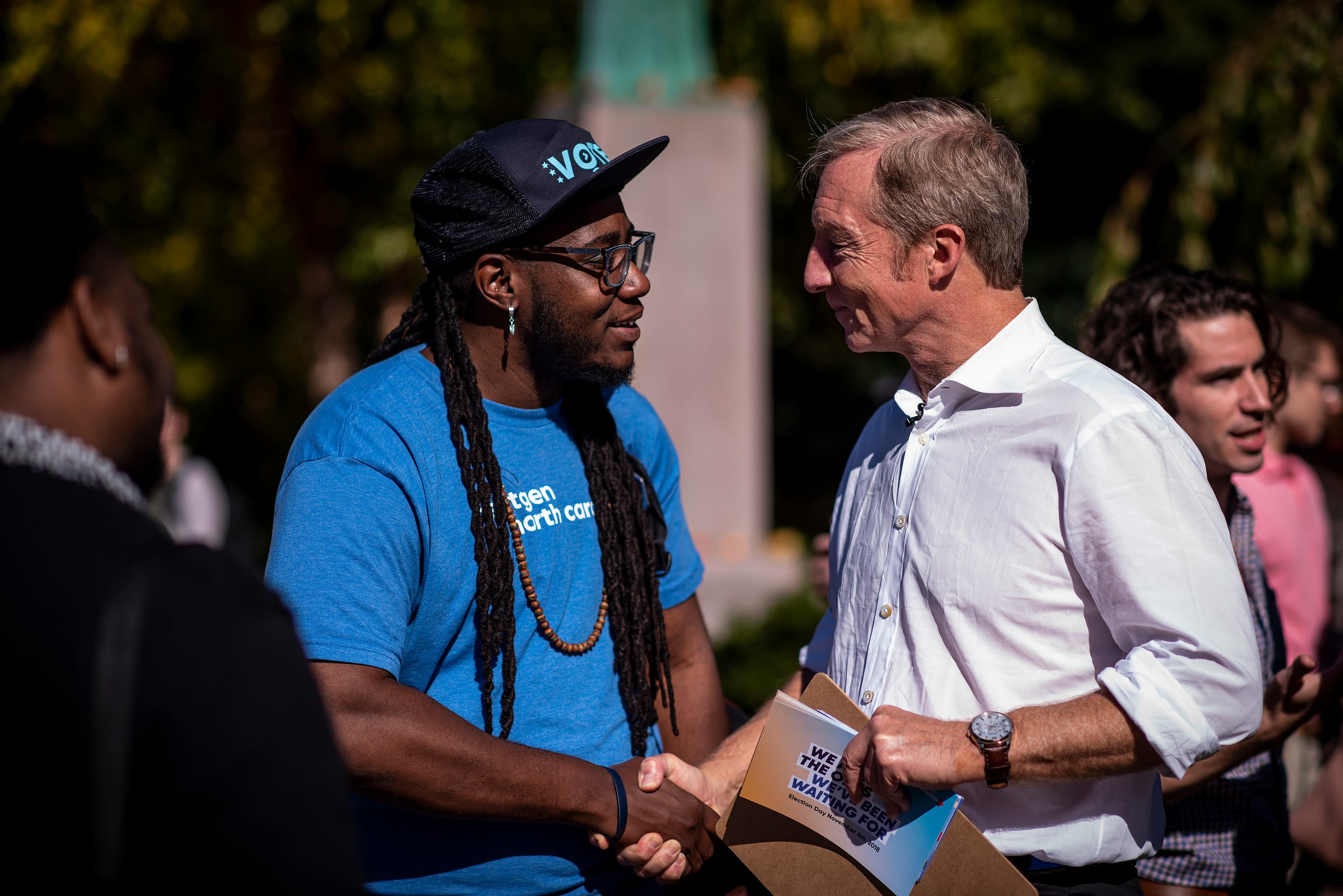 Two men shaking hands outdoors in a park. One wears a blue t-shirt and black cap, while the other wears a white button-up shirt. They are smiling and holding papers. There are other people blurred in the background, and trees surround them.