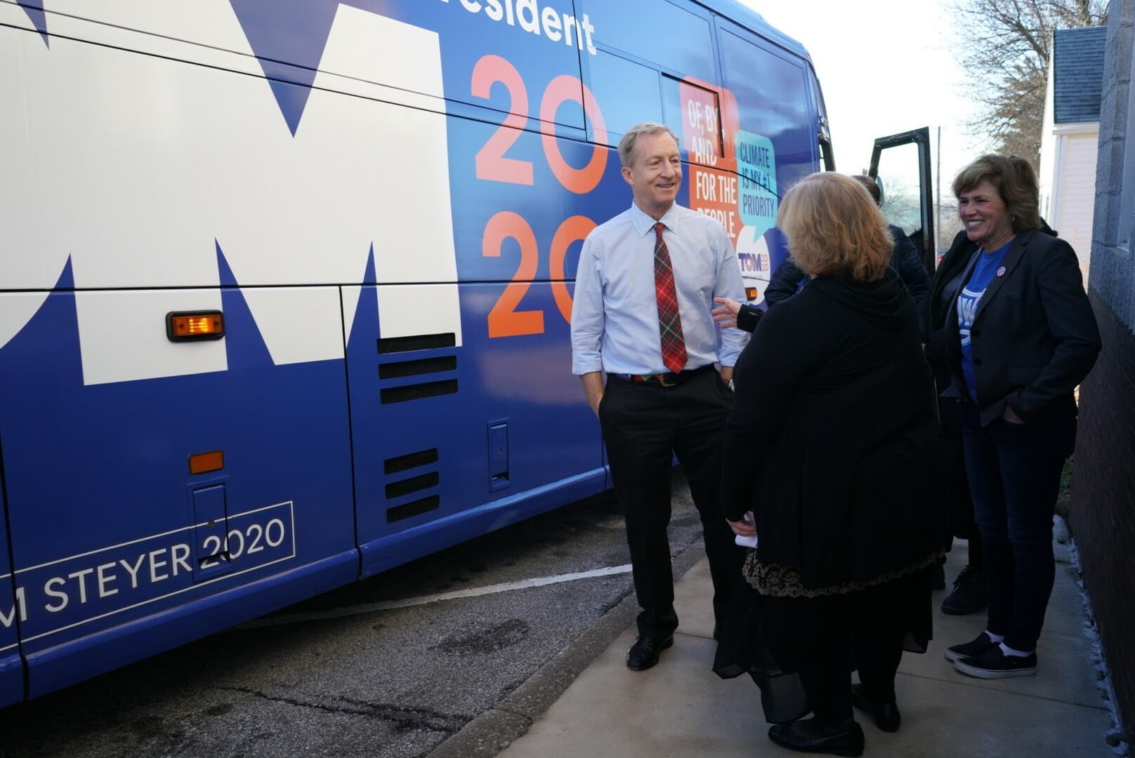 Three people standing in front of a blue campaign bus with "Tom Steyer 2020" and "2020" written in large letters. One man in a shirt and tie engages with two women, one wearing a jacket and the other in a coat. A "Climate Change is a Priority" sign is visible.