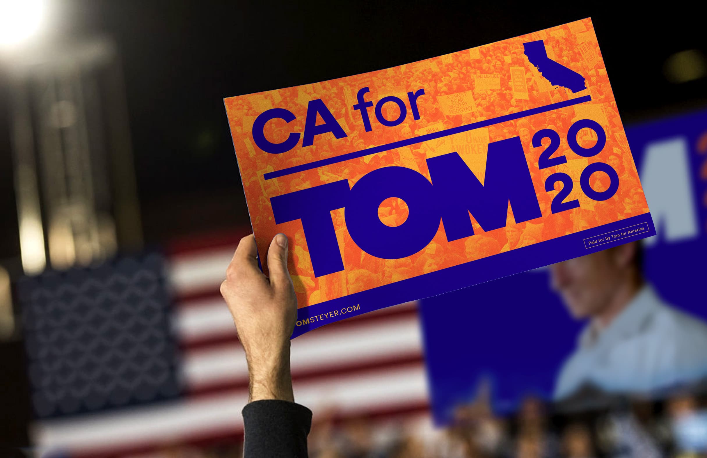 A person is holding up an orange sign that reads "CA for TOM 2020" in blue letters with a silhouette of California included in the design. The background is blurred, showing an American flag and a person speaking on stage.