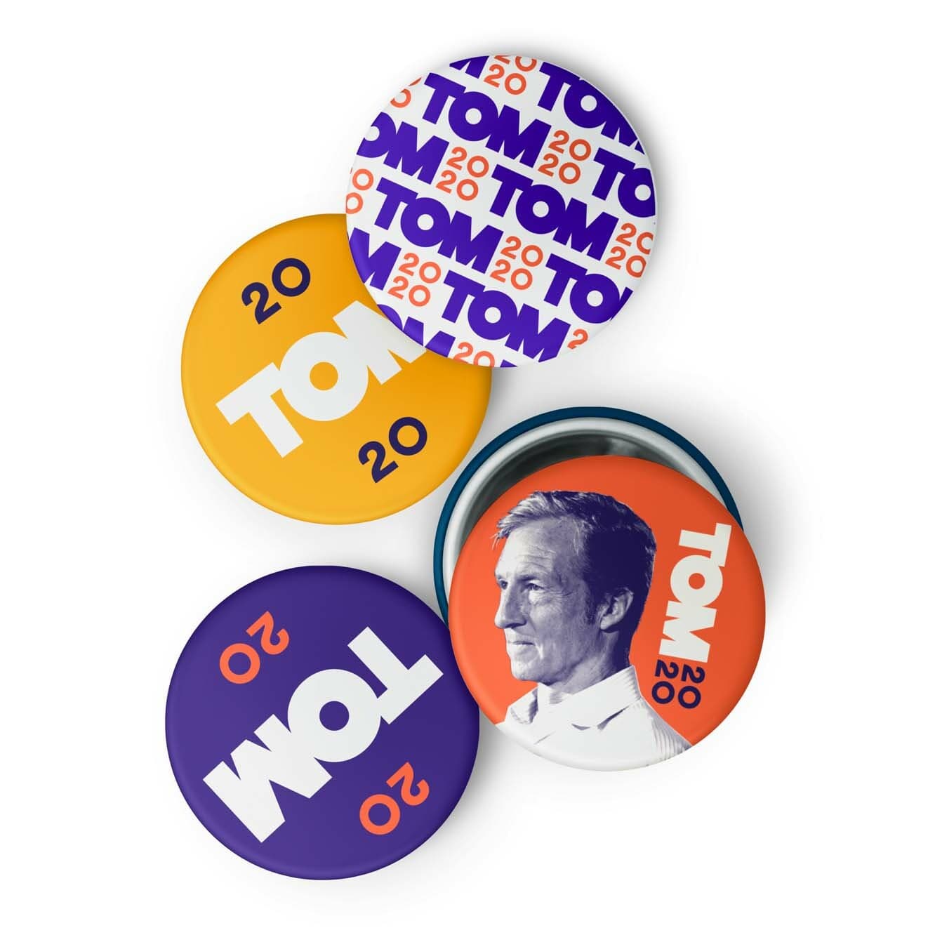 A close-up of four campaign buttons. Three buttons have text: "TOM 20," each in different styles and colors (purple, orange, and white backgrounds with contrasting text). One button has a black and white profile image of a man with the text "TOM 20" on an orange background.