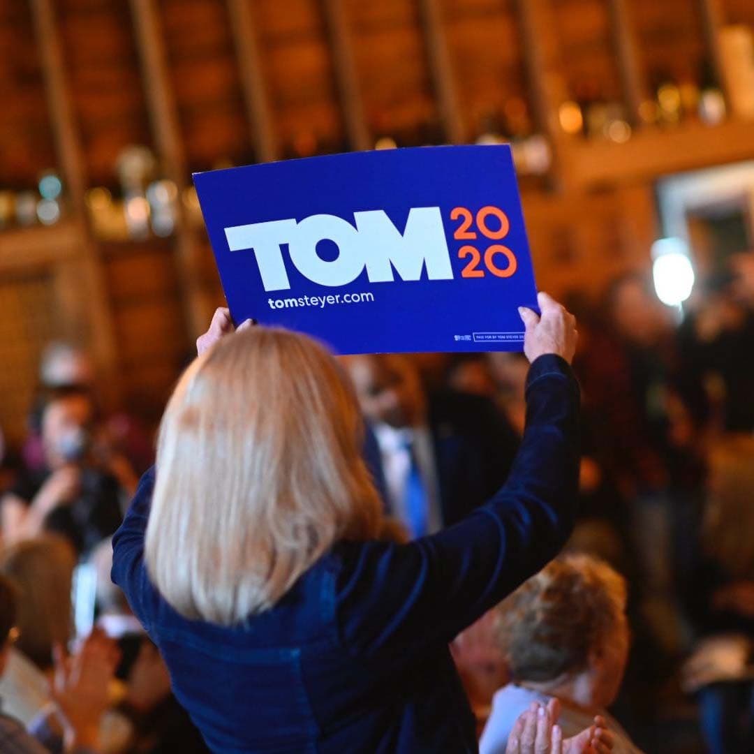 A person with blonde hair holds up a blue sign that reads "TOM 2020" in white and orange text. The background appears to be an indoor setting with a blurred audience and wooden walls.