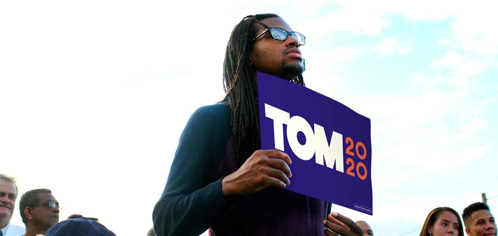 A person wearing glasses and with long hair stands outdoors holding a "TOM 2020" campaign sign. There are several people in the background, and the sky is clear.