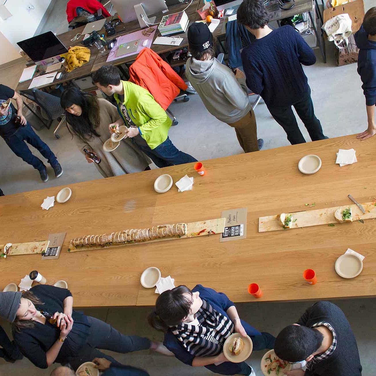 A group of people is gathered around a long wooden table with food and drinks, chatting and eating. The table has a long sandwich, plates, and bottles. Some people are standing while others are seated. The background shows desks and shelves in a room with a casual atmosphere.