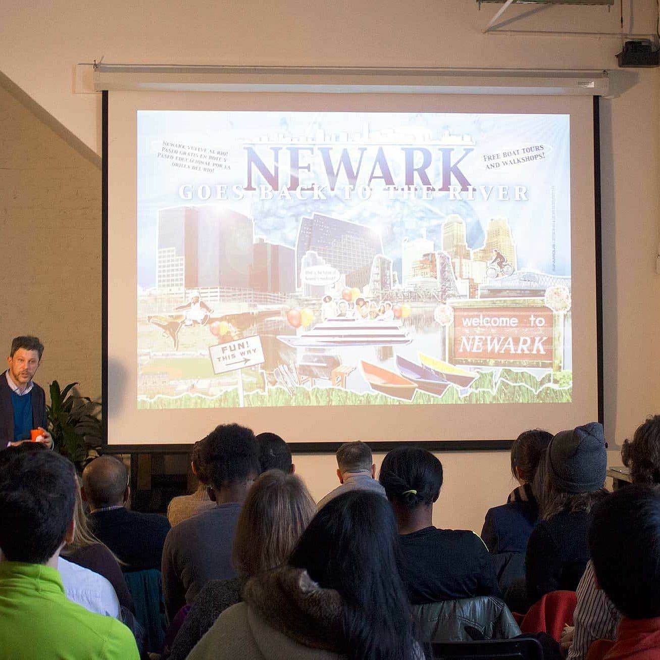 A man standing in front of an audience gives a presentation. He is near a large projection screen displaying a slide with the title "Newark Goes Back to the River" and images of cityscapes, a park, and welcome signs for Newark. Attendees are seated and attentive.