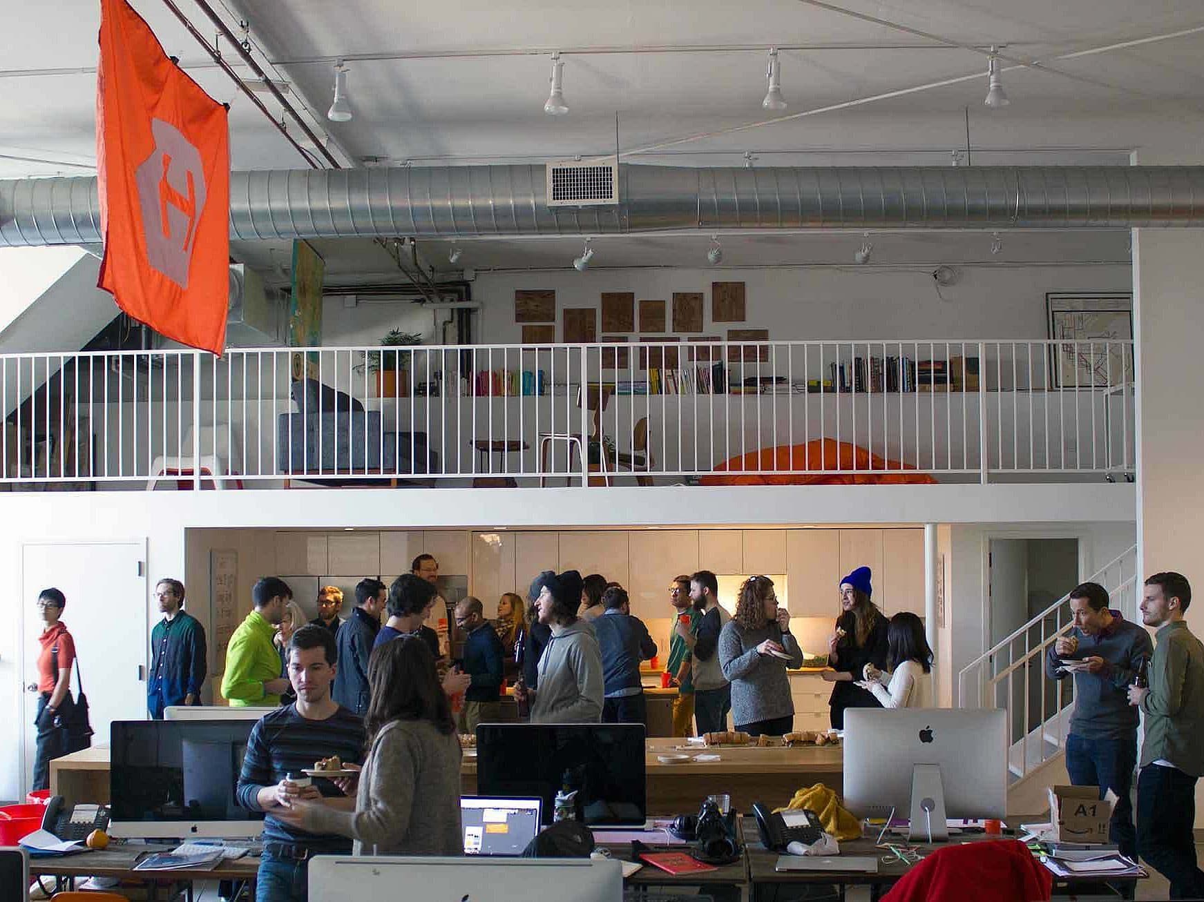 A group of people socializing in an open office space. The office has a mezzanine level and workstations on the ground floor. A large orange flag with a design hangs from the ceiling. Computers and office materials are visible on the desks.