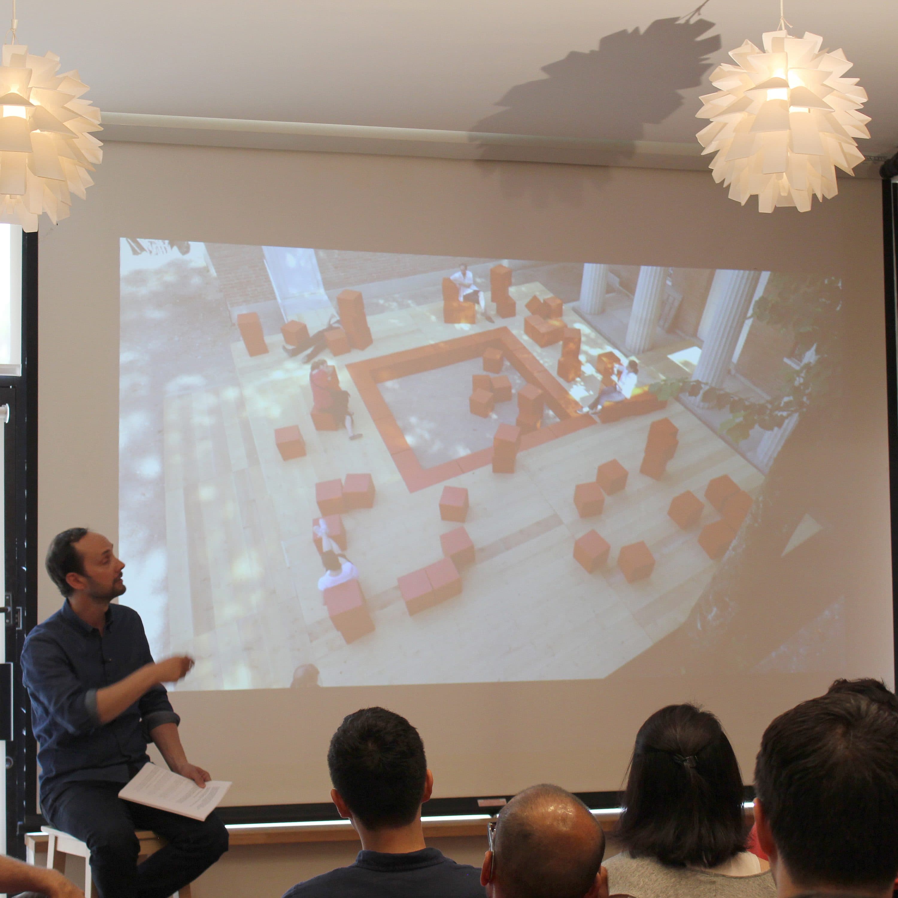 A man is presenting to an audience using a large screen. The screen displays an image of an outdoor scene with people interacting with brown block-like structures arranged in a square. The room has two decorative hanging lights and brick walls.