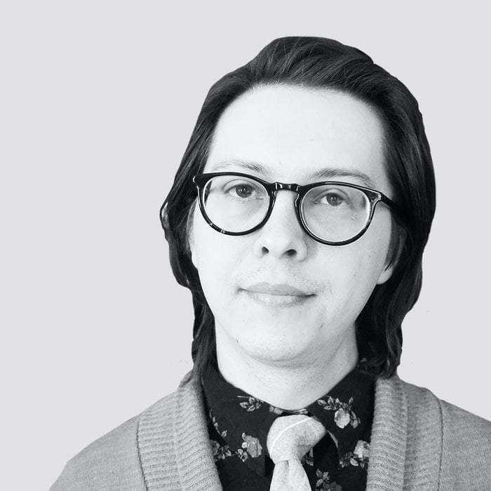 A person with shoulder-length dark hair and large, round glasses is pictured against a plain background. They are wearing a patterned shirt, a tie, and a cardigan. The photograph is in black and white.