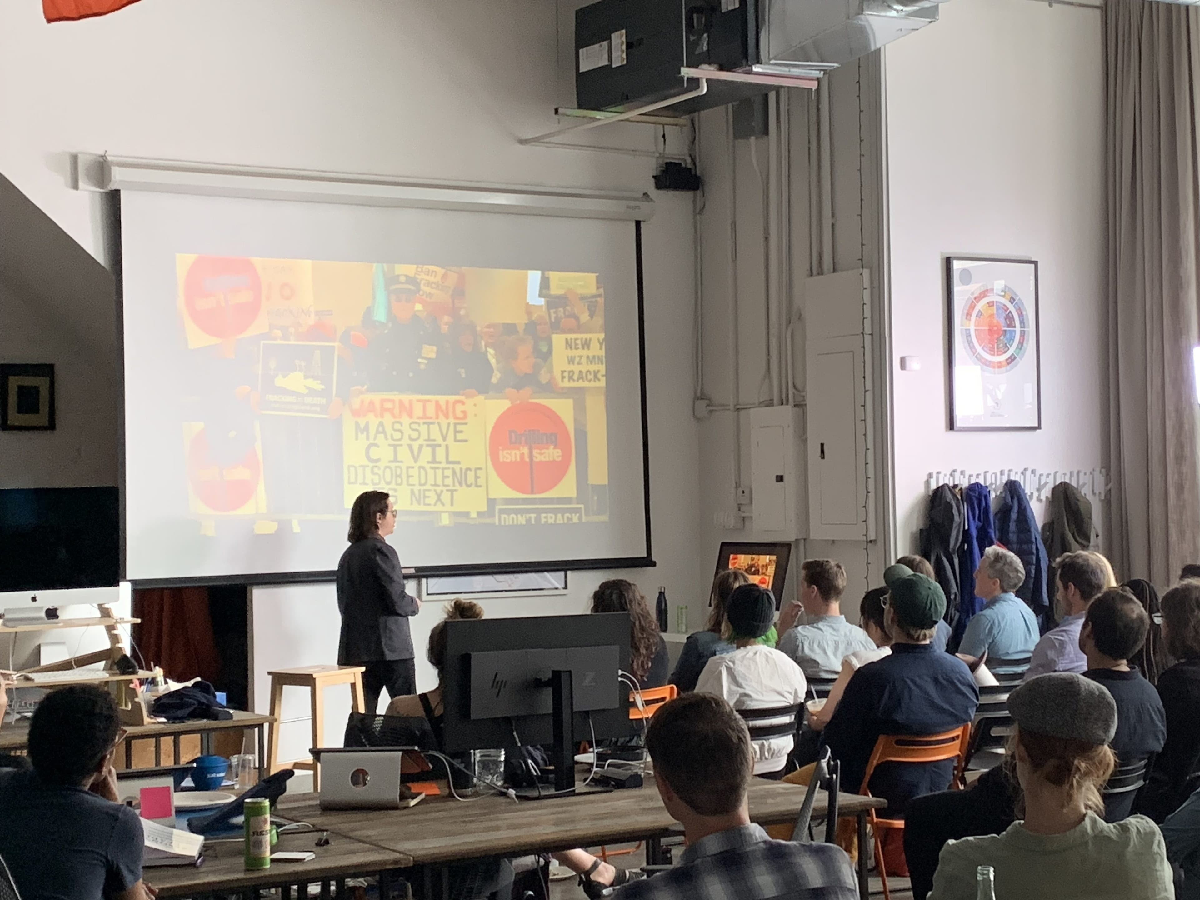 A group of people attentively listen to a person presenting in front of a projected screen. Various protest signs and images with messages like "Warning: Massive Civil Disobedience Next" are displayed. The setting appears to be a casual conference room or classroom.