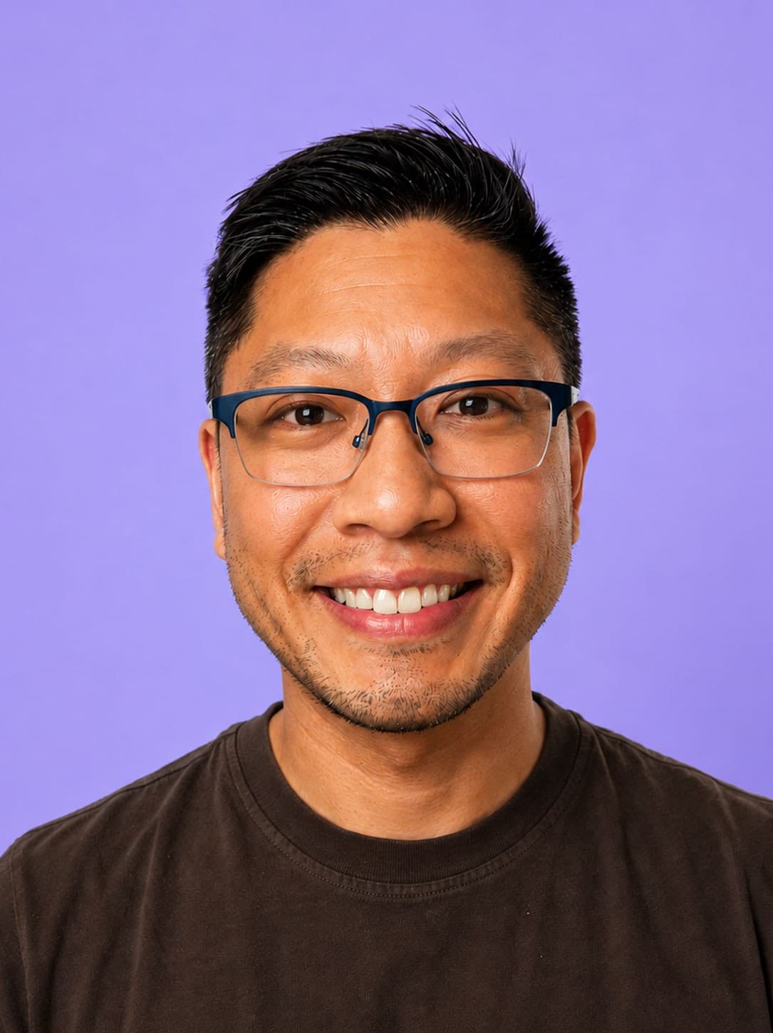 Smiling man with short dark hair, glasses, and a dark t-shirt stands in front of a solid light purple background.