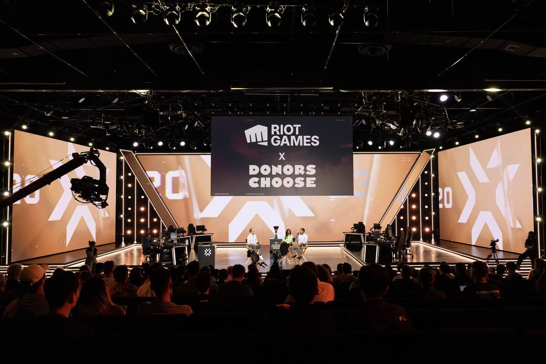 A live event stage with a large "Riot Games x Donors Choose" screen in the background. Several people are seated and engaged in conversation on stage. The audience sits facing the stage, and there are camera operators capturing the event.