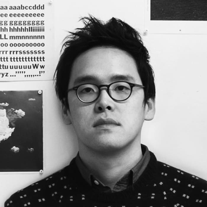 A person with short, tousled hair and round glasses stands against a wall. They are wearing a dark patterned sweater over a collared shirt. Behind them are various papers and images, including one with rows of repeated letters and punctuation marks. The photo is in black and white.