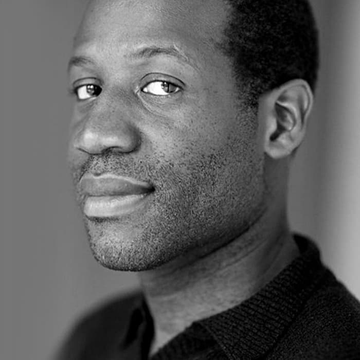 A person with short, curly hair and a slight stubble is looking at the camera with a neutral expression. They are wearing a dark-colored sweater, and the background is blurred. The image is in black and white.