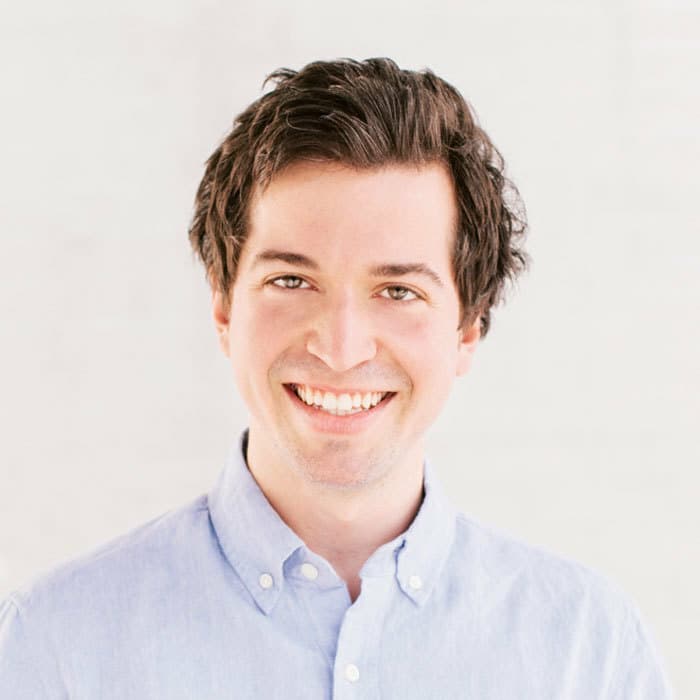 A person with short, dark hair is smiling at the camera. They are wearing a light blue button-up shirt and standing against a plain, light-colored background.