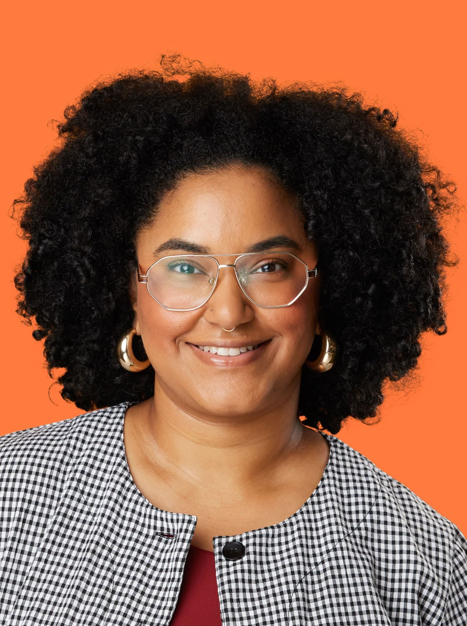 A person with curly hair, wearing glasses, gold hoop earrings, and a black-and-white checkered top, smiles in front of an orange background.