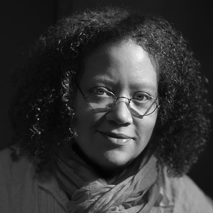 A black and white portrait of a person with curly hair and glasses, wearing a scarf and a cozy top. The lighting casts soft shadows, giving a warm and contemplative expression.