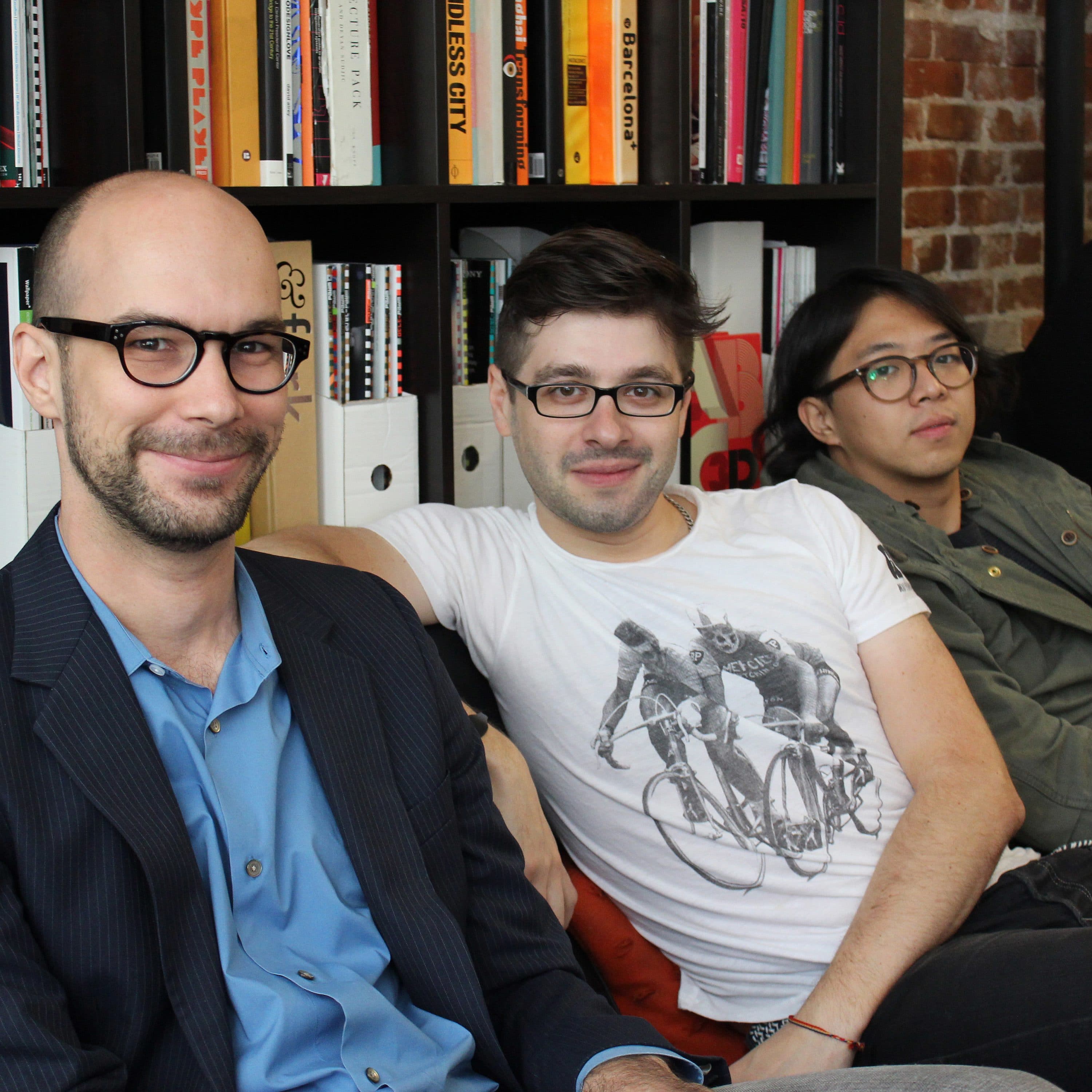 Three people are seated on a couch in front of shelves filled with books. The first person on the left wears glasses, a blue shirt, and a blazer, smiling at the camera. The second person, also wearing glasses, has on a white T-shirt with a cycling graphic. The third person, wearing a green jacket, looks relaxed with hands in pockets. Additional people in the background are partially visible.