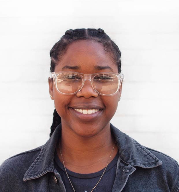 A person with braided hair, wearing clear glasses, a grey shirt, and a dark jacket, smiles warmly at the camera. The background is a plain, light-colored wall.