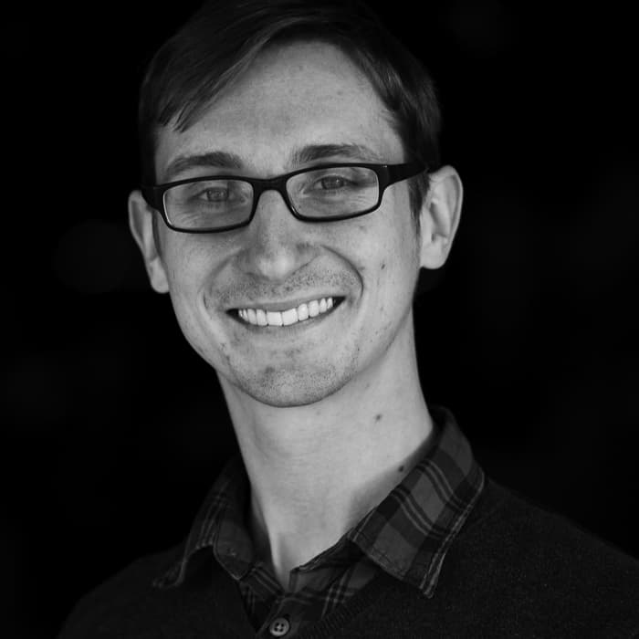 Black and white photo of a person wearing glasses and smiling. They have short hair and are dressed in a collared plaid shirt under a sweater. The background is dark, making the person the main focus of the image.