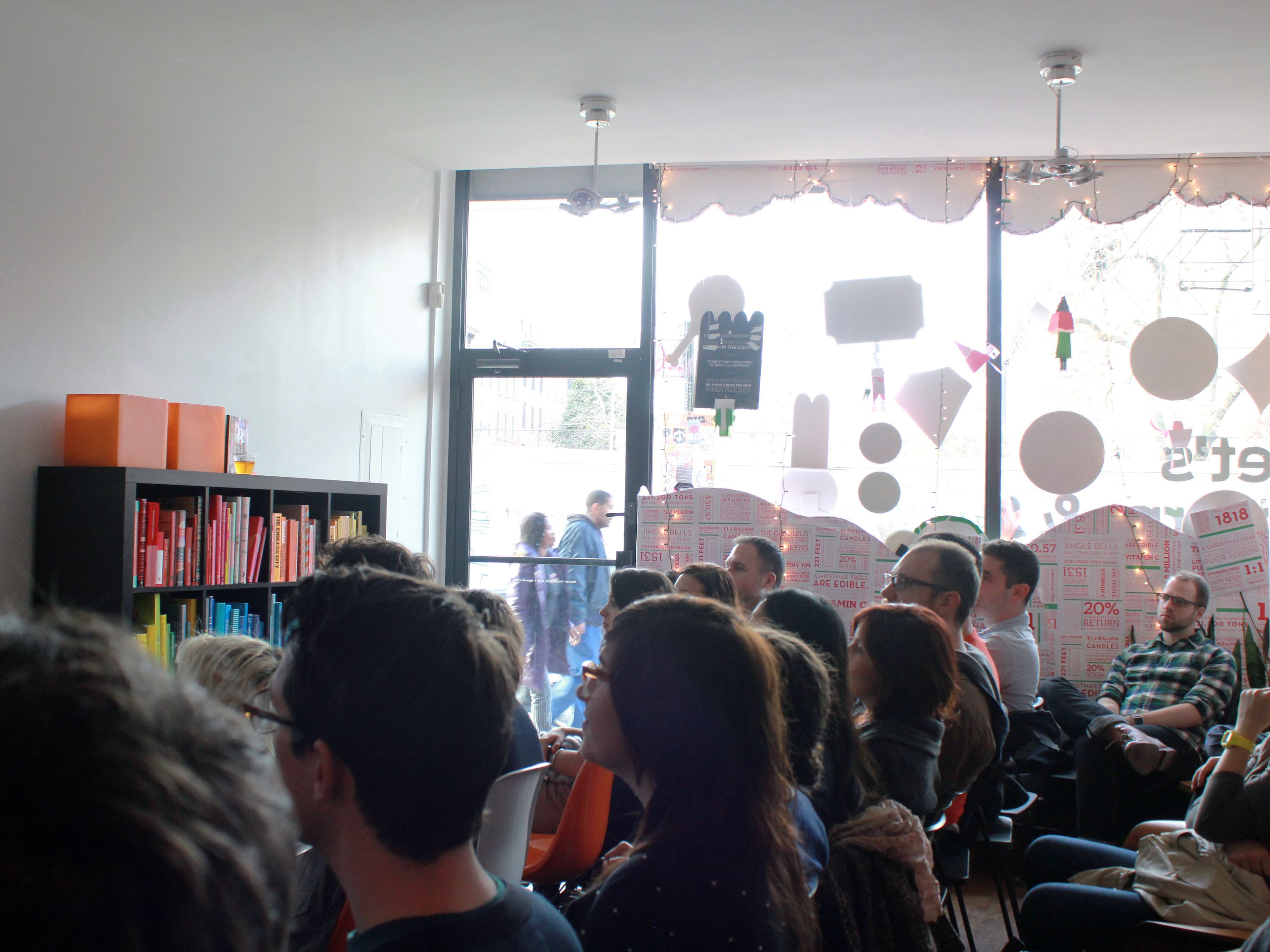 A group of people is seated in a small room, facing a bookshelf organized by color. The room has large windows letting in natural light, and people are visible outside. The windows are decorated with festive ornaments and promotional posters.