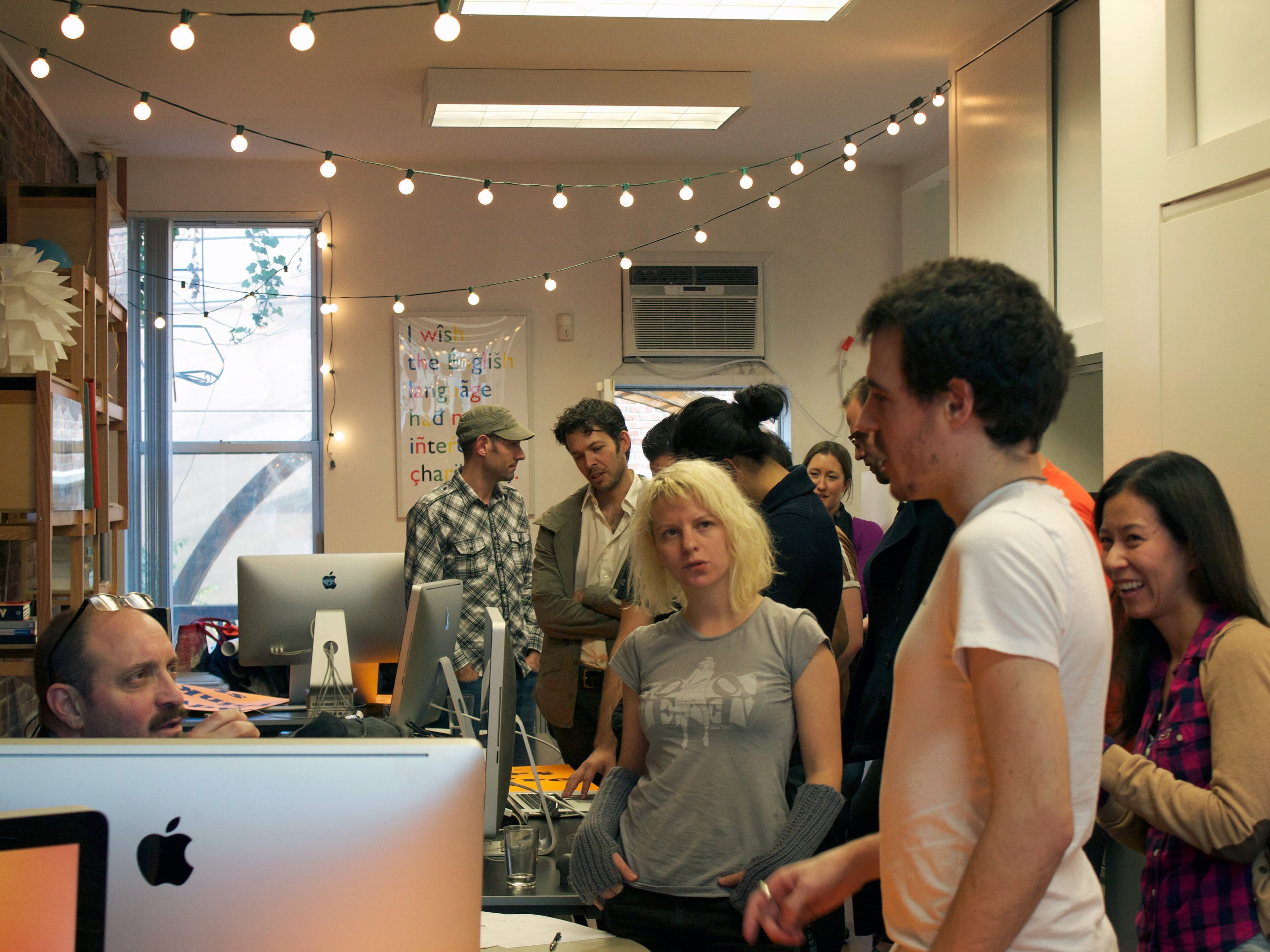 A lively office space with people mingling and working on computers. String lights hang from the ceiling, adding a festive touch. The atmosphere is casual, with individuals chatting, looking at screens, and enjoying the social environment.