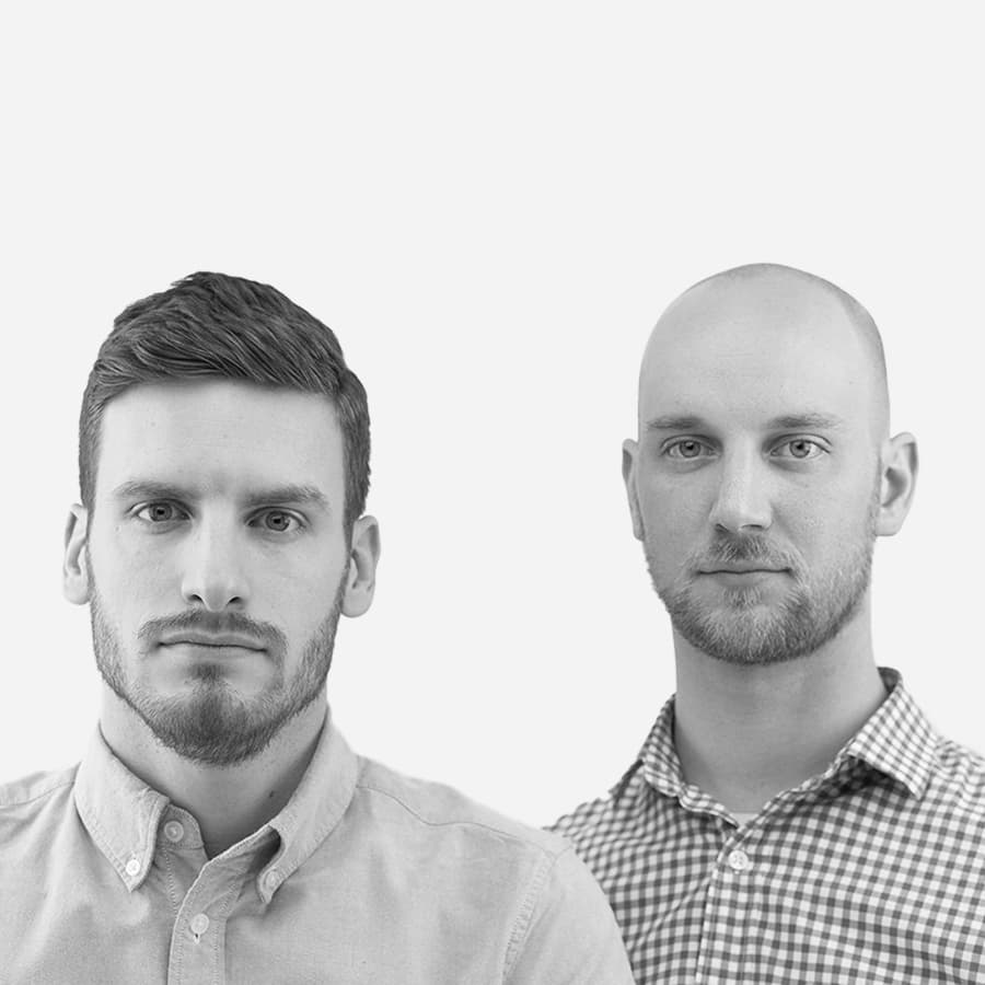 A black and white image featuring two men. The man on the left has short dark hair and a beard and is wearing a light-colored button-up shirt. The man on the right is bald with a beard and is wearing a checkered button-up shirt. They are both looking directly at the camera.