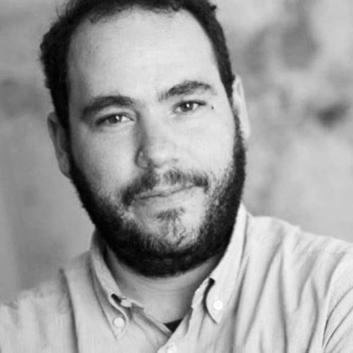 A man with a beard and short hair, wearing a button-up shirt, is looking into the camera with a slight smile. The image is in black and white, with a blurred background that appears to be a textured wall.