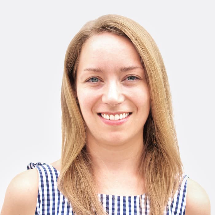 A person with long light brown hair is smiling at the camera. They are wearing a sleeveless, blue and white checkered top. The background is plain white.