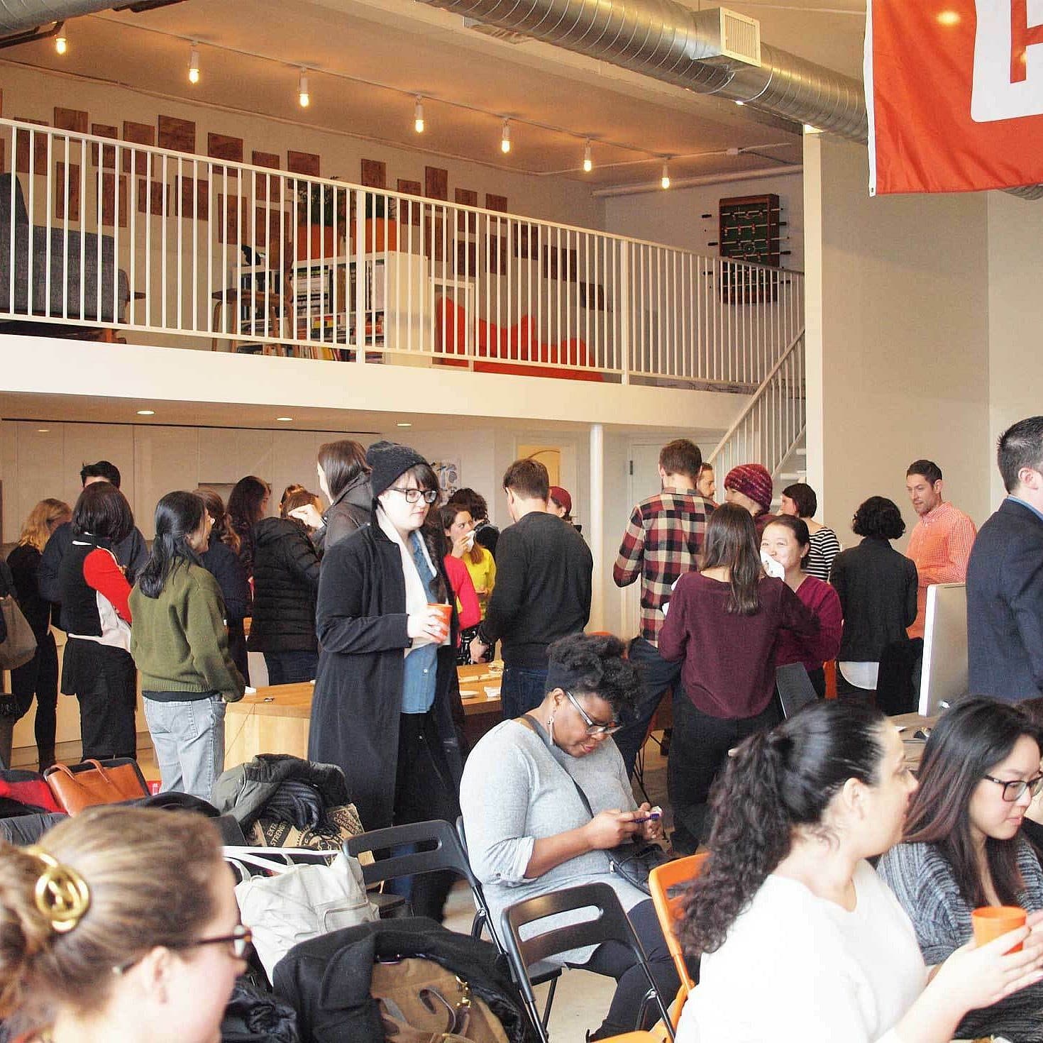 A diverse group of people gather in a spacious room with a high ceiling and a mezzanine. Some people stand and chat, while others sit and eat. The room is well-lit by string lights, and an orange flag with a white logo hangs on the wall.