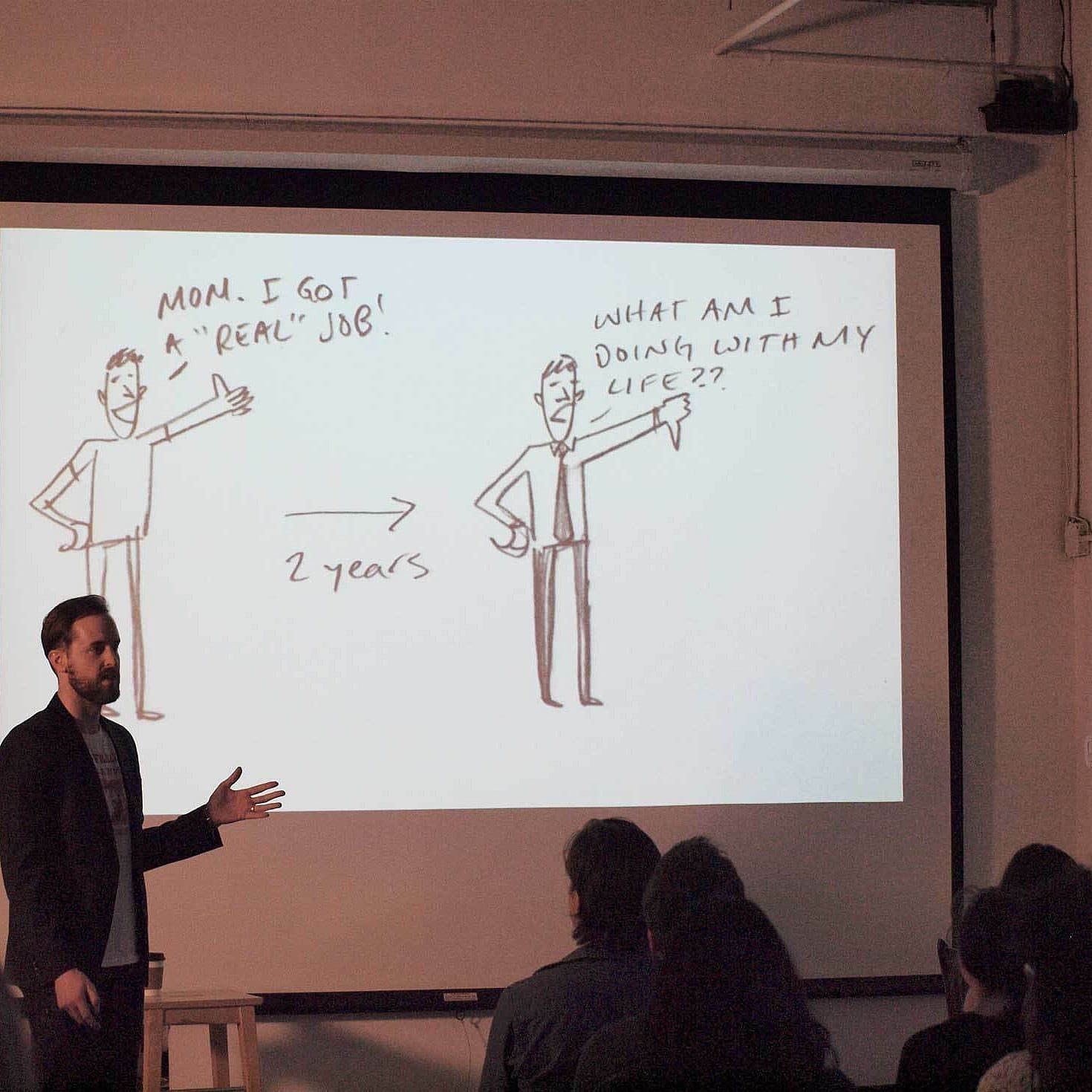 A presenter stands before an audience, pointing at a projected illustration. The slide shows two cartoon figures: one saying "Mom, I got a REAL job!" with an arrow pointing to another figure asking, "What am I doing with my life??" The time span "2 years" is noted.