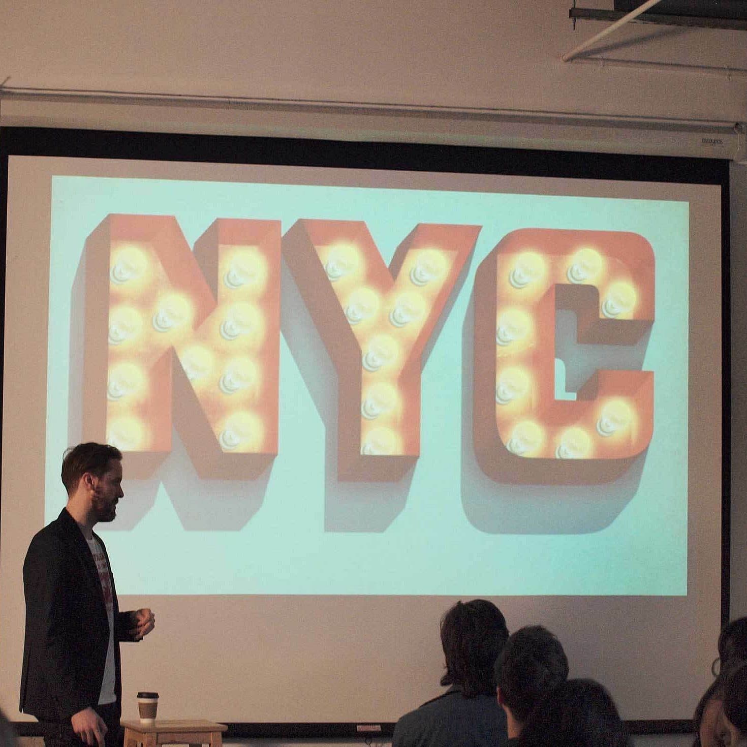 A man stands in front of an audience presenting a slide with large, illuminated letters spelling "NYC." The room is dimly lit, and the audience is facing the presenter and the screen, which dominates the background.