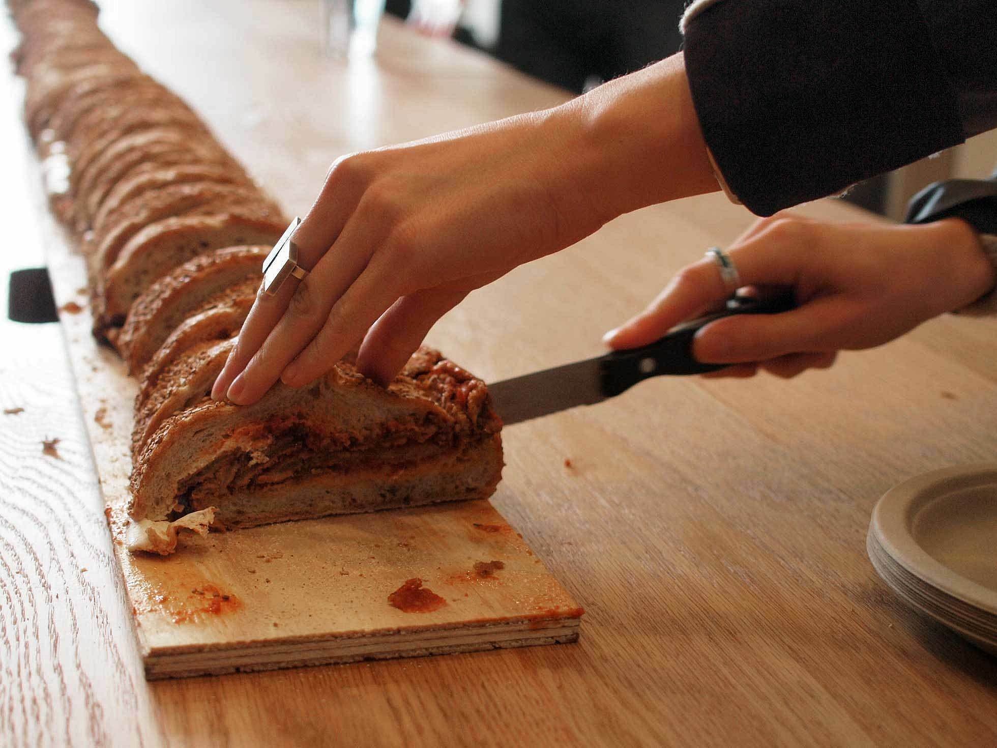 A person's hands are cutting a long loaf of bread with a knife on a wooden cutting board. The bread appears to have a filling, possibly tomato or a similar spread. The person has rings on their fingers. A plate is visible next to the cutting board.