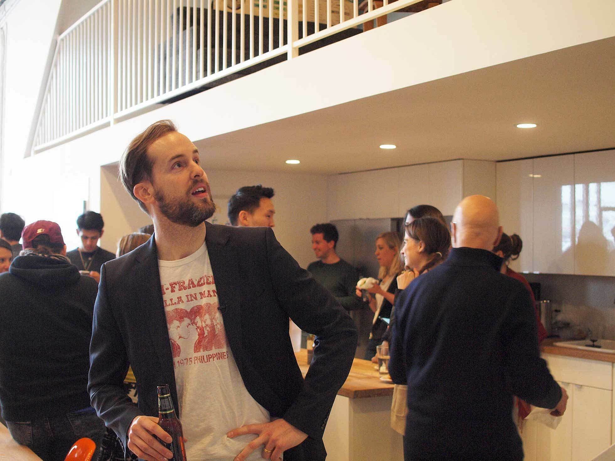 A man with a beard, wearing a T-shirt and dark blazer, holds a drink while looking upwards in a bustling kitchen. Several people are gathered in the background, engaged in conversation and activities. White cabinets and a loft area are visible in the modern kitchen.