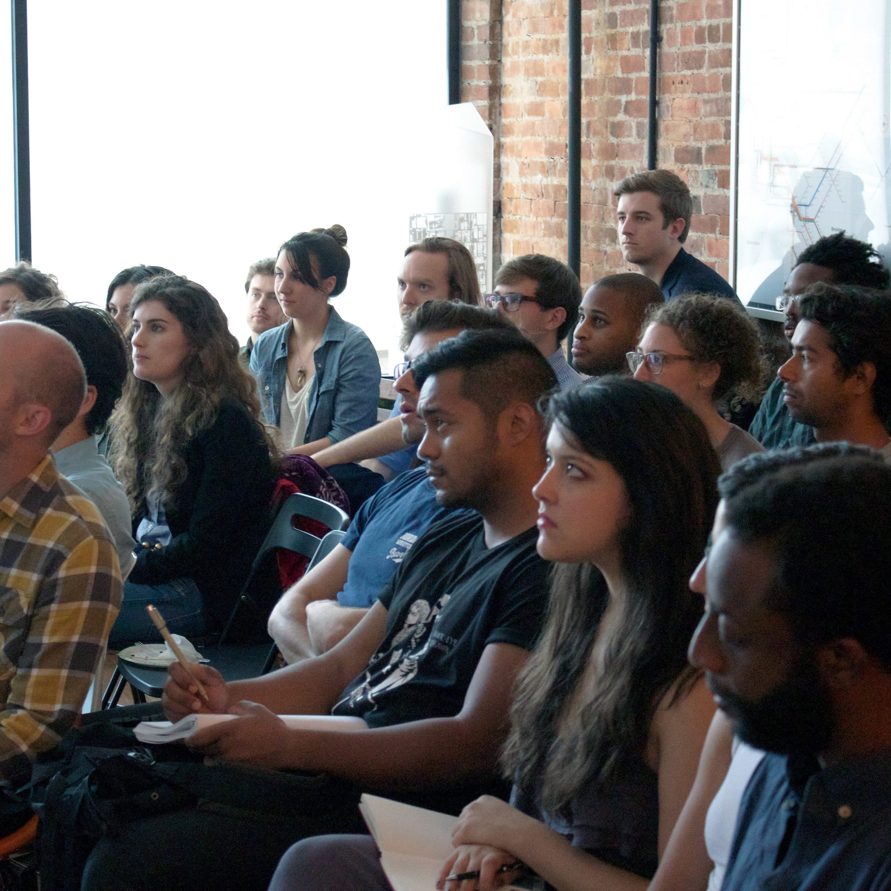 A group of people seated and attentively listening to a presentation in a room with exposed brick walls. Some are taking notes while others are looking towards the front. The room is well-lit with natural light coming through a large window.