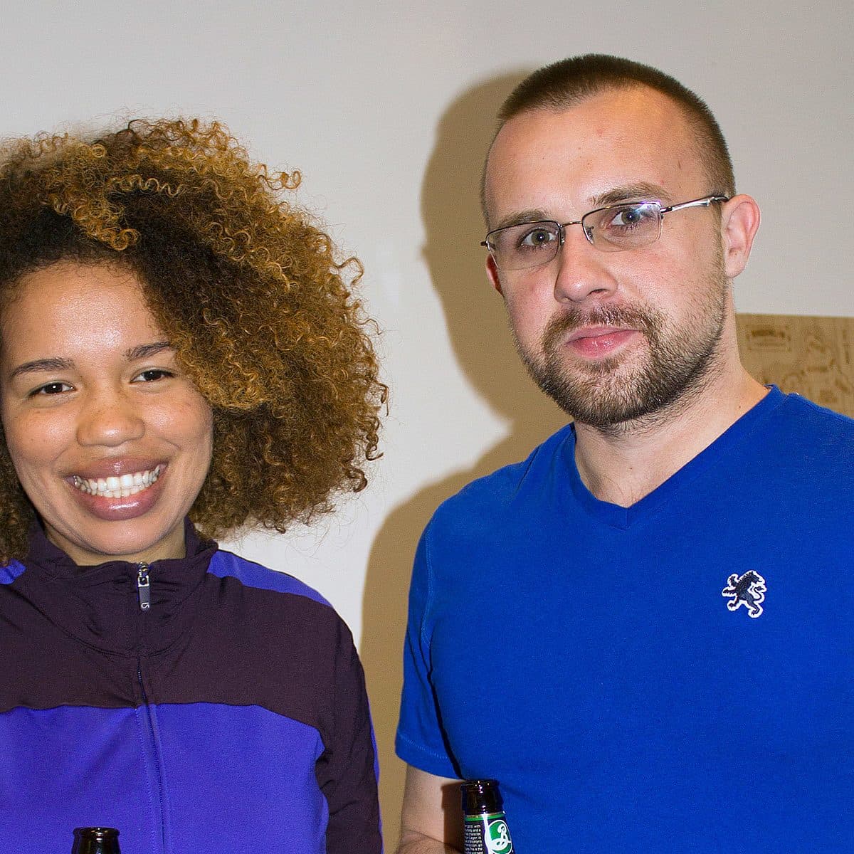 A woman with curly hair wearing a purple jacket and a man with glasses and a beard wearing a blue shirt are both smiling and holding beer bottles. They are standing against a light-colored wall with some artwork in the background.