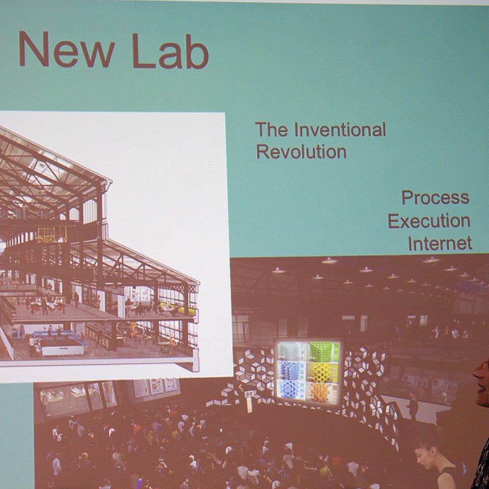 A woman stands in front of a presentation slide titled "New Lab". The slide features images of a large industrial warehouse and a busy workshop. Text on the slide includes "The Inventional Revolution," "Process," "Execution," and "Internet.
