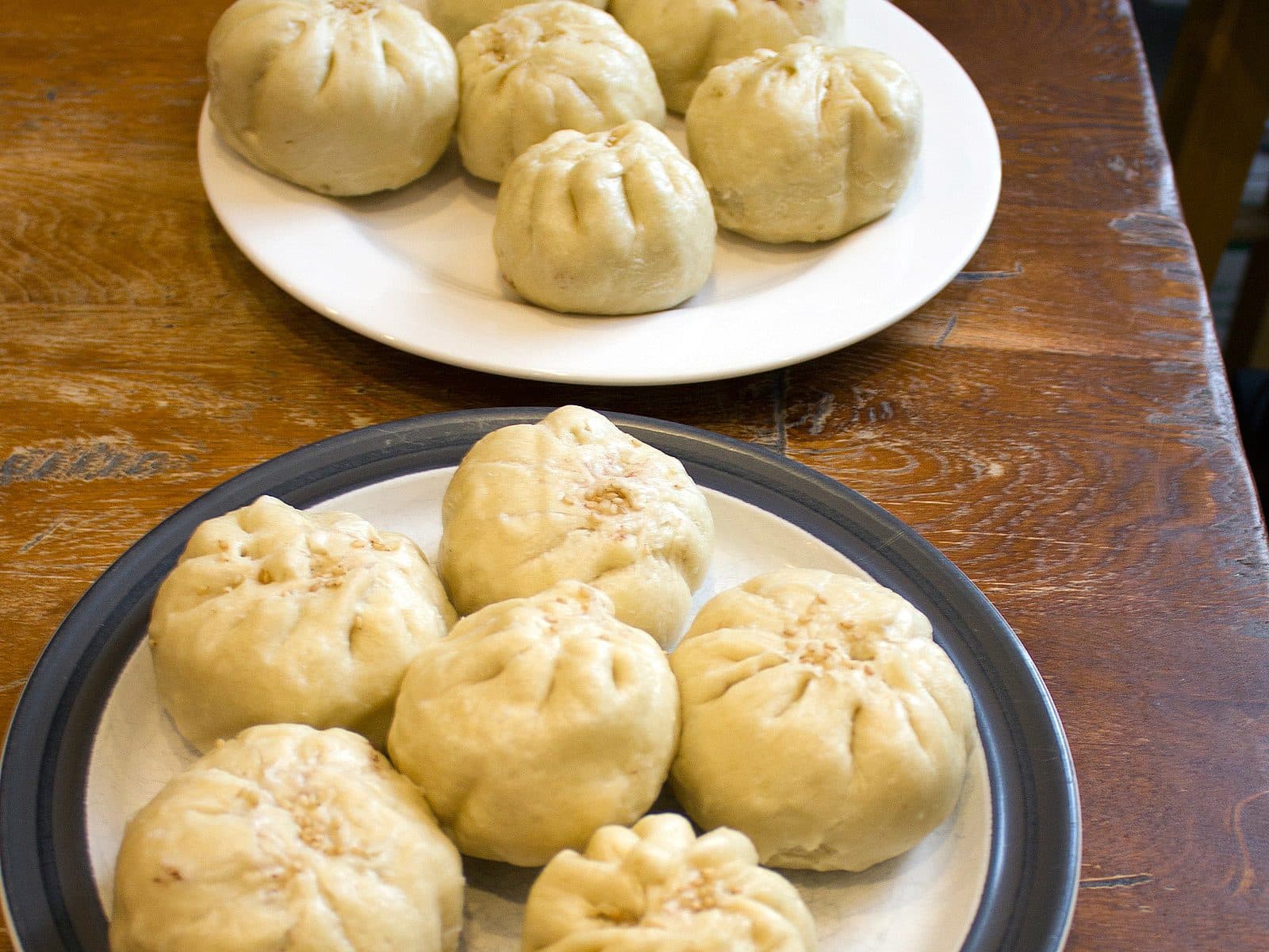 Two plates of freshly steamed buns on a wooden table. The front plate is black and white, holding six buns. The back plate is white with six buns as well. Each bun is pinched at the top, showcasing a soft, fluffy texture.