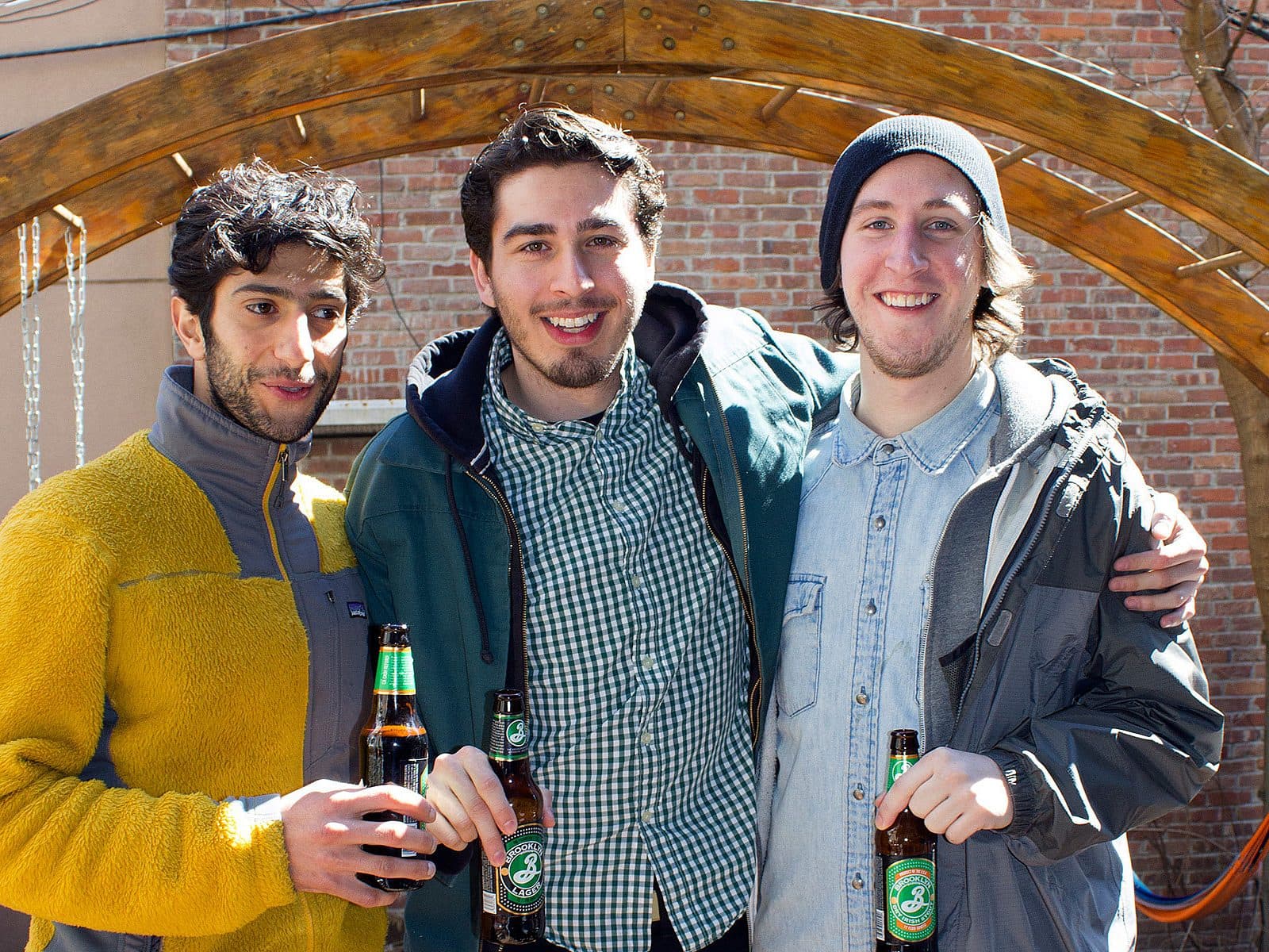 Three young men stand closely together outdoors, all smiling at the camera. They are each holding a beer bottle. The man on the left wears a yellow and gray jacket, the middle one a green jacket and plaid shirt, and the one on the right a gray beanie and jacket. A wooden arch is in the background.