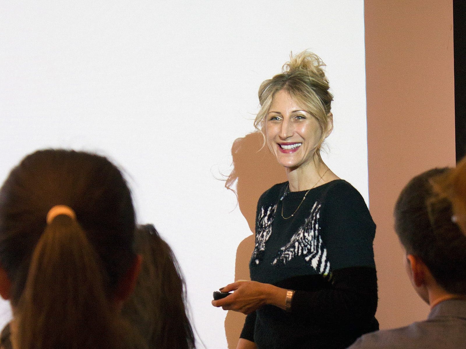 A woman with blonde hair in a bun and wearing a black sweater is standing in front of a projection screen, smiling and holding a presentation remote. She is facing a seated audience whose backs are visible in the foreground.