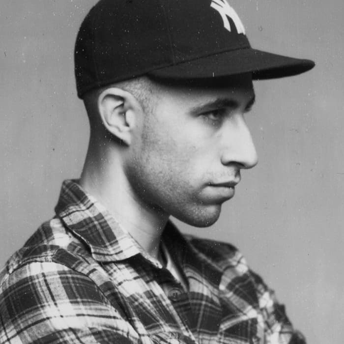 A black-and-white side profile photograph of a person wearing a checkered flannel shirt and a baseball cap. The individual has a neutral expression and is looking to the right. The background is plain and untextured.