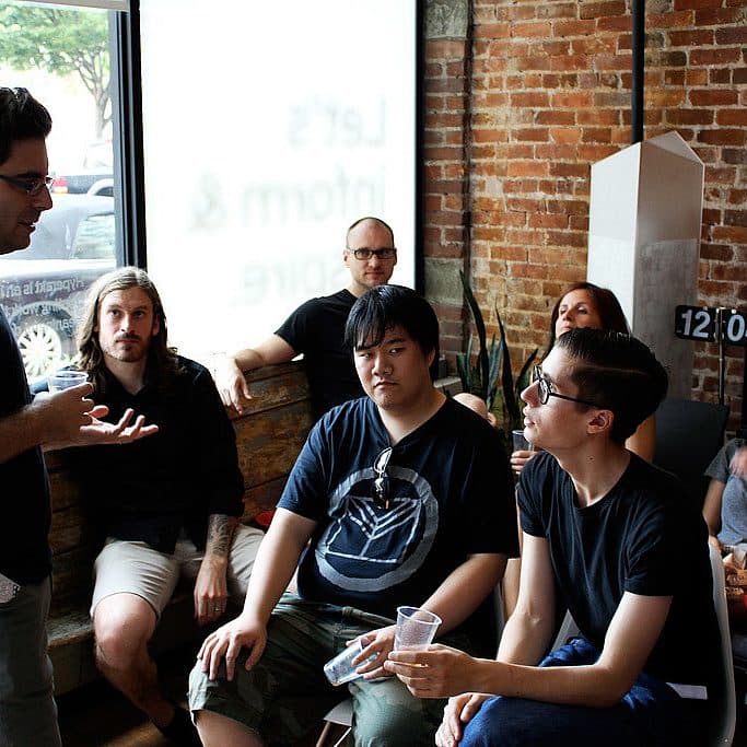 A group of people sits and stands in a casual discussion in a room with brick walls and large windows. One person stands and speaks, while others sit on a wooden bench and chairs, some holding plates and cups. The atmosphere is relaxed and informal.