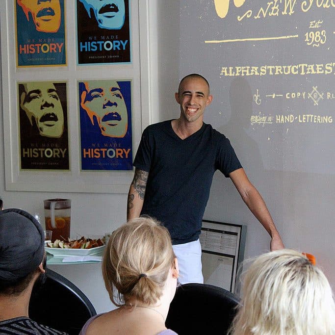 A man stands smiling in front of a group of seated people, giving a presentation. He is next to a screen displaying text and has a whiteboard behind him. The room has posters with a graphic design of a face and text reading "We Made History" on the wall.