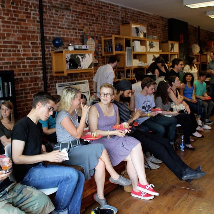 A group of people is seated closely together on benches in a room with exposed brick walls, enjoying food and drinks. Bookshelves and various objects are visible in the background, and a few individuals are standing and conversing. The atmosphere appears casual and social.