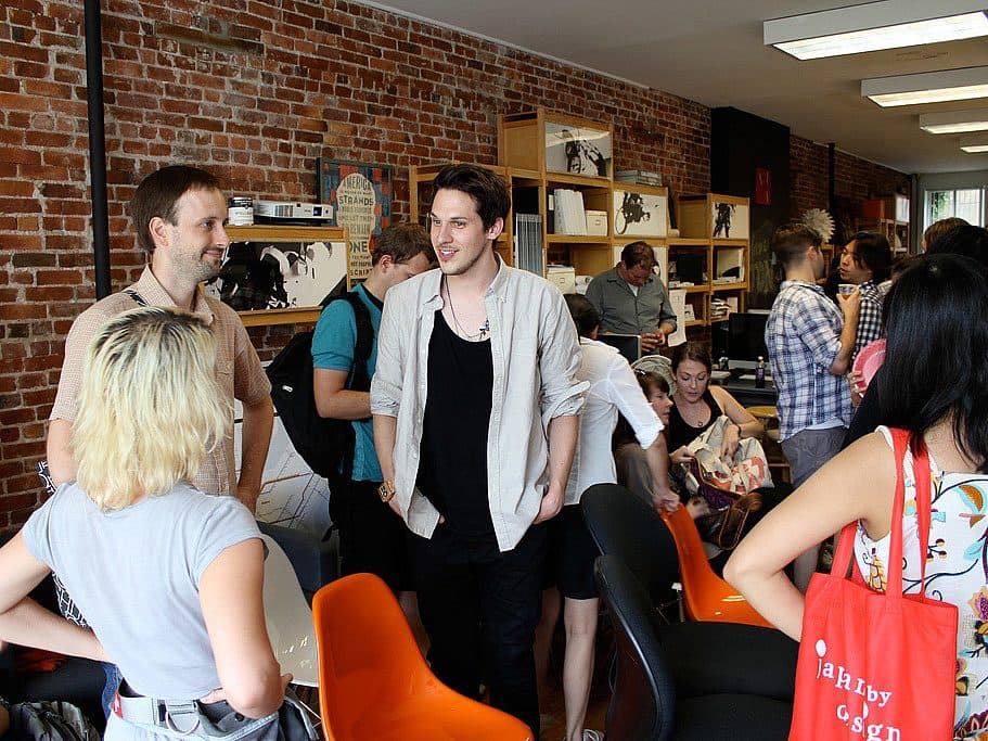 A group of people socializing in a room with brick walls. Some are standing around, while others are seated on colorful chairs. Shelves with various items are in the background, and everyone appears to be engaged in conversation and enjoying the gathering.