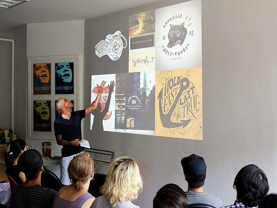 A presenter gestures towards a wall with various graphic design images projected on it, while an audience sits and watches. The designs include logos, typography, and illustrations. The room has posters on the left wall showing the word "HISTORY".